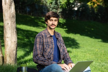 Bıyıklı, genç, sakallı, esmer bir öğrenci parkta dizüstü bilgisayarla oturmuş kameraya bakıyor. Uzak bir yerden çalış. Parkta serbest çalışan biri.