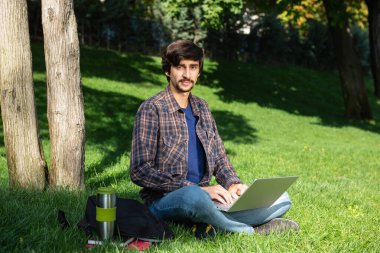 Bıyıklı, genç, sakallı, esmer bir öğrenci parkta dizüstü bilgisayarla oturmuş kameraya bakıyor. Uzak bir yerden çalış. Parkta serbest çalışan biri.