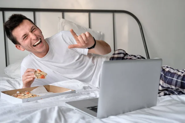 Young man in the white shirt and pajama smiling lying on a bed and eating tasty pizza. Concept of food delivery. Staying home during quarantine.