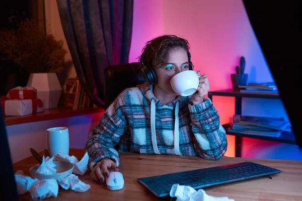 Young woman with eyeglasses and wireless headphones watching video on a computer and drinking coffee from a cup. Mess and used napkins on a table. Neon light at the background. Work from home.