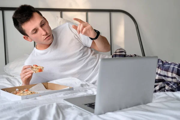 Young man in the white shirt and pajama watching video on laptop, cheering for the sports team lying on a bed and eating tasty pizza. Concept of food delivery. Staying home during quarantine.