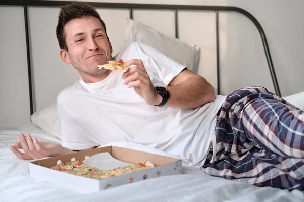 Young man in the white shirt and pajama smiling lying on a bed and eating tasty pizza. Concept of food delivery