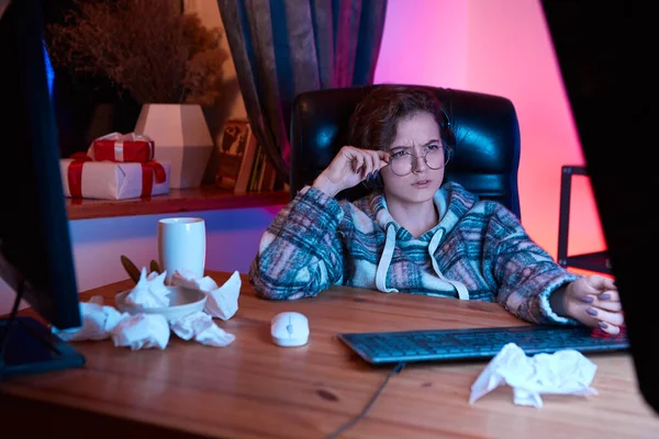 Confused young woman with eyeglasses and wireless headphones looking at the screen of a monitor. Mess and used napkins on a table. Neon light at the background. Work from home