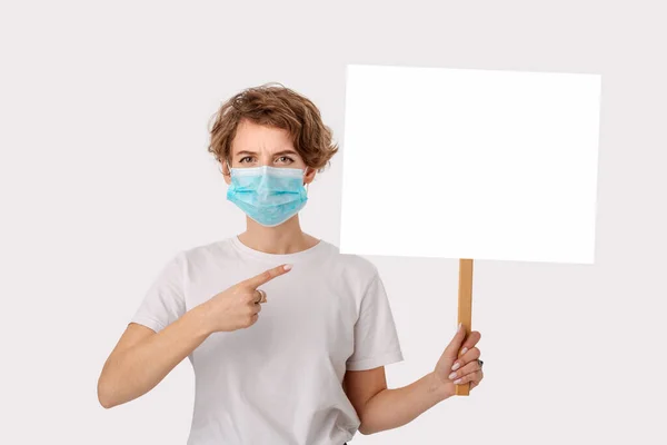 青い医療用マスクをした白いシャツを着た若い女性が ピケットのサインのコピースペースを指差しているのを心配している 保護病のことを考えて ロイヤリティフリーのストック写真