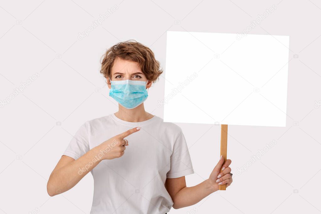 Worried young woman in a white shirt wearing blue medical mask holds pointing at the copy space of a picket sign. Think about protection disease.