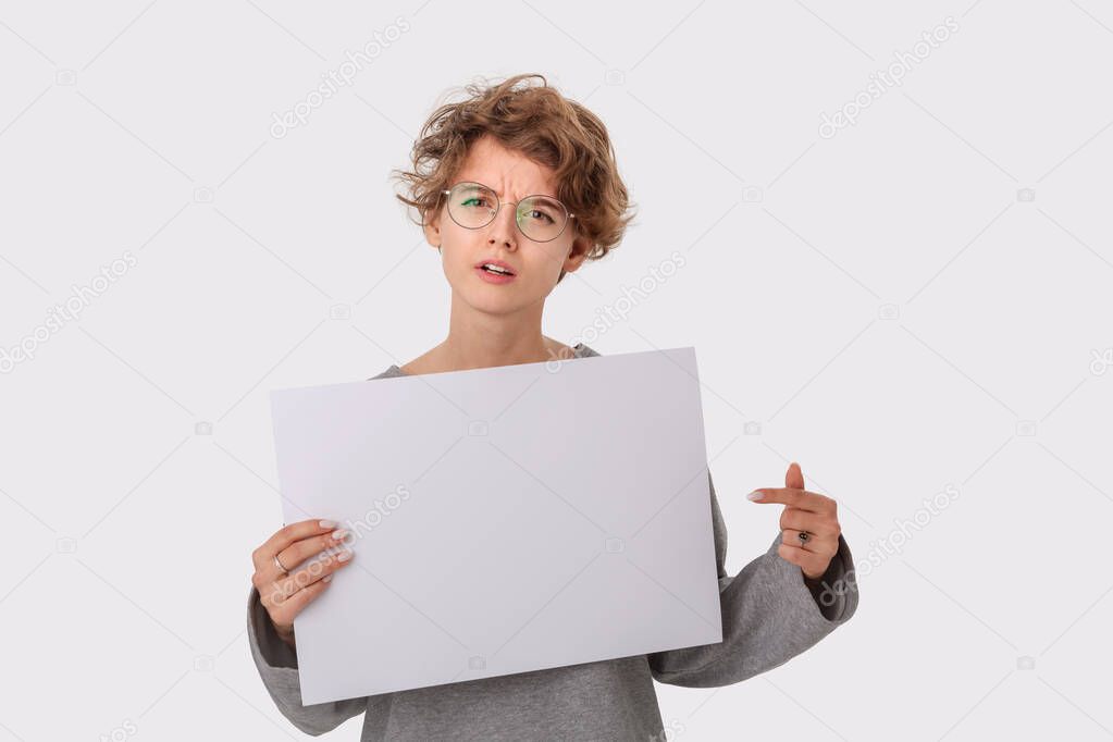 Concerned young woman with eyeglass holding empty blank paper board with copy space for text and pointing at it with finger. Copy space on paper