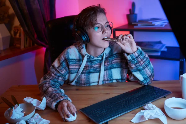 Pretty young woman with eyeglasses and wireless headphones watching video on a computer and eating chocolate. Neon light at the background. Work from home during isolation