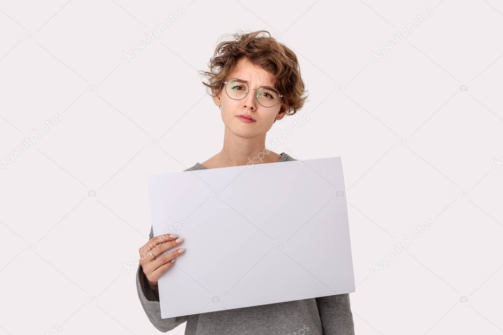 Concerned young woman with eyeglass holding empty blank paper board with copy space for text and pointing at it with finger. Copy space on paper