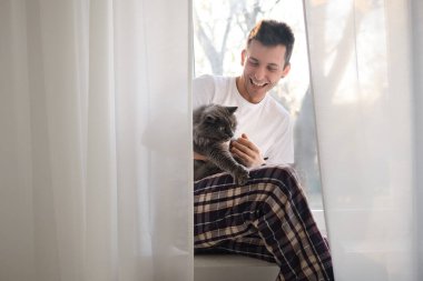 Beyaz gömlekli ve pijamalı genç adam gri tüylü kediyi okşuyor. Evcil hayvanlarla hayat. İzolasyon sırasında boş zaman aktiviteleri