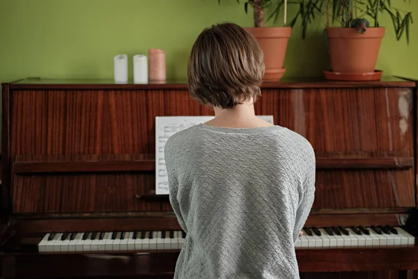 自宅でアコースティックピアノを弾いている若い女性のバックビュー ホーム イゾレーション 自宅で快適に感じている ピアノの植木鉢の花 隔離生活 ロイヤリティフリーのストック画像