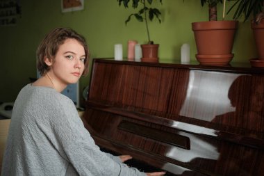 Akustik piyanonun yanında oturan kameraya bakıp gülümseyen genç bir kadın. Ev adası. Evde rahat hissediyorum. Piyanodaki saksılarda çiçekler. Karantinada yaşam tarzı.