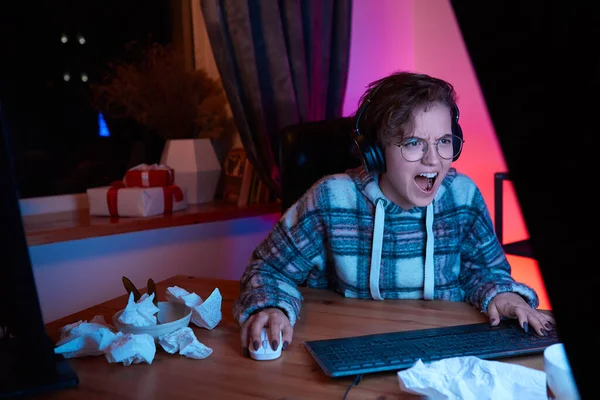 Angry young woman with eyeglasses and wireless headphones shouting at monitor. Mess and used napkins on a table. Neon light at the background. Work from home. Isolation during quarantine.