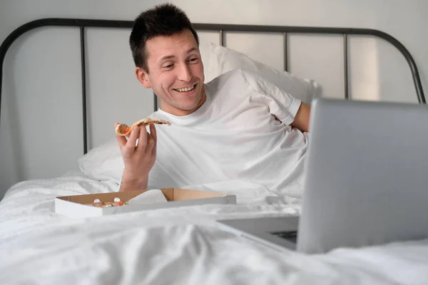 Young man in the white shirt and pajama watching video on laptop lying on a bed and eating tasty pizza. Concept of food delivery. Staying home during quarantine.