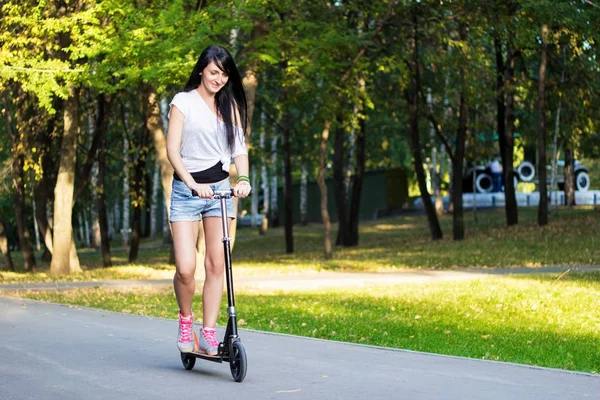 Vista de cerca de atleta morena jinete femenino en zapatos grises y pantalones cortos está montando una patada scooter en carretera de asfalto en el parque en la tarde soleada verano — Foto de Stock