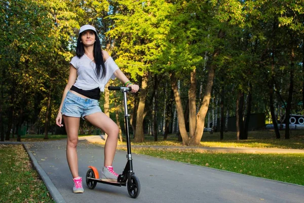 Atleta morena adolescente femenina está de pie cerca de patada scooter en el parque en verano noche soleada —  Fotos de Stock