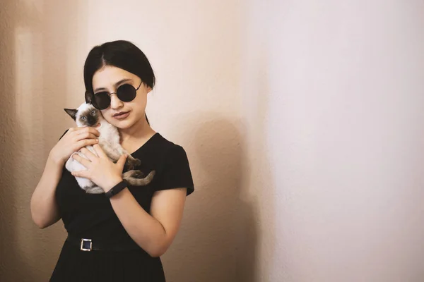 Jovem mulher atraente abraçando bichano gato em mãos. Menina bonito e glamouroso na moda óculos de sol posando com seu gato siamês — Fotografia de Stock