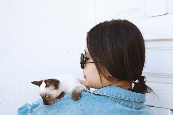 Joven mujer atractiva abrazando coño gato en las manos. Linda y glamorosa chica en gafas de sol de moda posando con su gato siamés — Foto de Stock