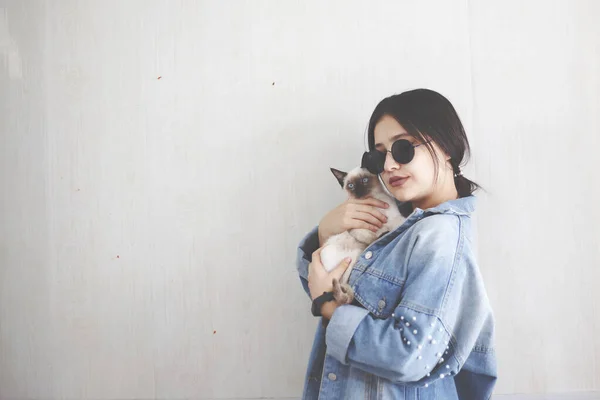 Jovem mulher atraente abraçando bichano gato em mãos. Menina bonito e glamouroso na moda óculos de sol posando com seu gato siamês — Fotografia de Stock