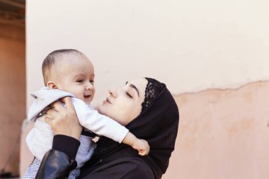 Bir yaşındaki bebeğini kucaklayan, sarılan ve öpüşen tesettürlü genç Müslüman kadın. Abaya giysili mutlu Müslüman anne. Öp ve küçük çocuğuyla oyna.