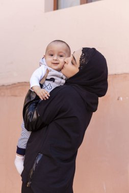 Bir yaşındaki bebeğini kucaklayan, sarılan ve öpüşen tesettürlü genç Müslüman kadın. Abaya giysili mutlu Müslüman anne. Öp ve küçük çocuğuyla oyna.