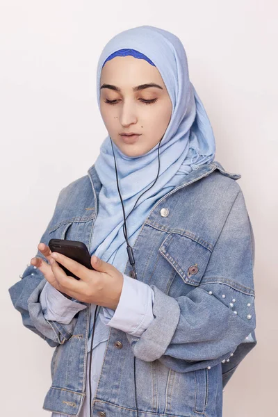 Menina muçulmana moderna ouve música, podcasts e audiolivros no smartphone. Mulher muçulmana feliz no hijab ouvindo músicas com fone de ouvido — Fotografia de Stock