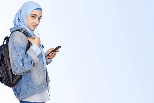 现代穆斯林女学生在智能手机上听音乐、播客和有声读物。 戴头巾的快乐的穆斯林女人用耳机听着歌曲 — 图库照片