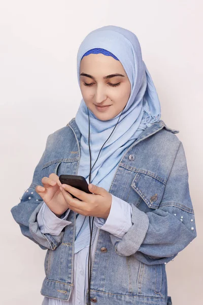 현대의 모슬렘 소녀는 스마트폰으로 음악, 팟캐스트, 오디오 북을 듣는다. 히잡을 쓴 행복 한 회교도 여인 이 헤드폰으로 노래를 듣고 있다 — 스톡 사진