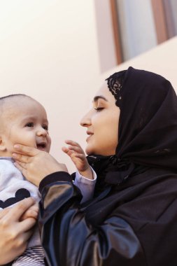 Bir yaşındaki bebeğini kucaklayan, sarılan ve öpüşen tesettürlü genç Müslüman kadın. Abaya giysili mutlu Müslüman anne. Öp ve küçük çocuğuyla oyna.