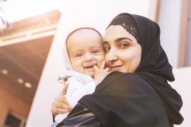 Bir yaşındaki bebeğini kucaklayan, sarılan ve öpüşen tesettürlü genç Müslüman kadın. Abaya giysili mutlu Müslüman anne. Öp ve küçük çocuğuyla oyna.
