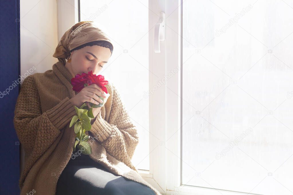 Young Muslim woman sits on the window sill with flower bouquet in hands. Depressed middle-eastern female near the window at home. Lonely cheerless girl feeling stressed and waiting
