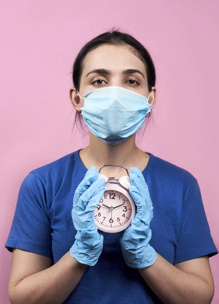 Young woman with a face mask and medical gloves holding an alarm clock. Coronavirus or Covid-19 pandemic. Quarantine time. Time to stay home during virus infection spread