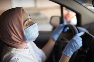 Cerrahi maske ve eldiven takmış genç bir Müslüman kadın Coronavirus salgını sırasında arabayı sürüyor. Tesettürlü ve koruyucu giysili araba kullanan kadın bir doktor. Kadın doktor işe gidiyor.