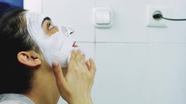 Banyoda makyaj maskesi takan bir kadın. Karışık ırk dişileri yüzüne beyaz besleyici maske ya da krem sürerler. Yüzüne fırça maskesi takıyorum. Cilt temizleme spa rahatlama konsepti