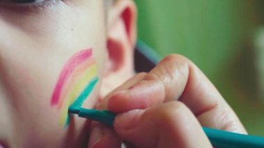Küçük bir çocuğun yakın çekim görüntüsü. LGBTQ ebeveyni yanaklarına gökkuşağı bayrağı çiziyor. Onur Günü 2020, LGBT ailesi, cinsiyet hakları ve eşitlik kavramları