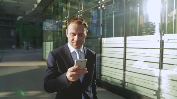 Aantrekkelijke zakenman met behulp van de app op smartphone, wandelen in de stad. Steadicam schot. — Stockvideo