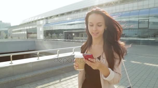 Hübsches Mädchen zu Fuß und per App auf dem Smartphone Kaffee trinken in der Sonne. Steadicam-Aufnahme. — Stockvideo