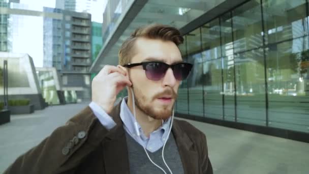 Homem atraente colocando fones de ouvido, ouvindo música, andando, dançando, bebendo café de uma xícara de papel — Vídeo de Stock