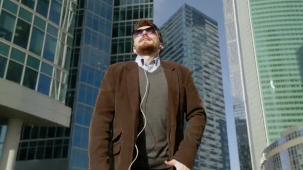 Hombre guapo escuchando música en auriculares para teléfonos inteligentes y bailando cerca de rascacielos — Vídeo de stock