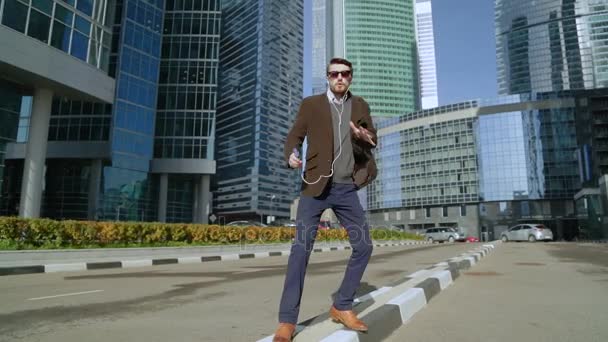 Красивый мужчина слушает музыку в наушниках и танцует возле небоскребов — стоковое видео