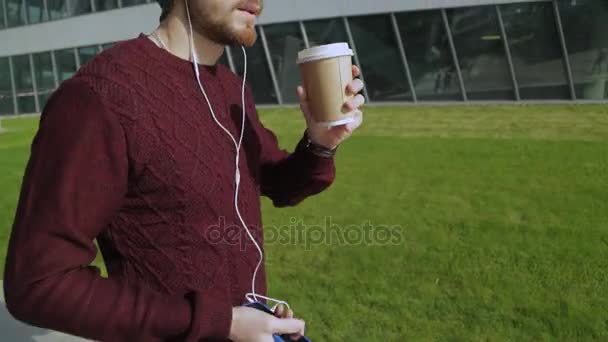 हँडसम हिप्स्टर स्मार्टफोन हेडफोन्समध्ये संगीत ऐकणे आणि कागदावरून कॉफी पिणे, चालणे आणि नृत्य — स्टॉक व्हिडिओ