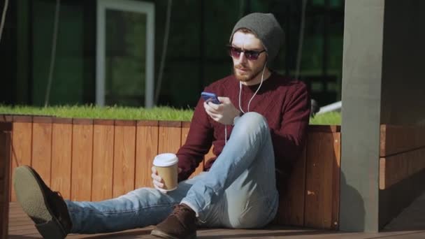 在智能手机耳机听音乐，喝咖啡从纸的英俊时髦 — 图库视频影像