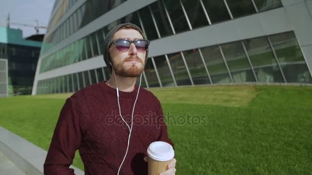 Guapo hipster escuchando música en auriculares para teléfonos inteligentes y bebiendo café de papel, caminando y bailando — Vídeos de Stock