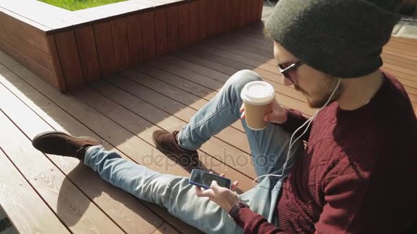 Guapo hipster utilizando la aplicación y escuchar música en los auriculares del teléfono inteligente — Vídeos de Stock