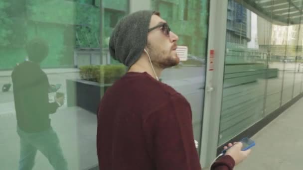 Knappe hipster met behulp van de app en luisteren naar muziek in smartphone hoofdtelefoon, wandelen en op zoek naar iets — Stockvideo