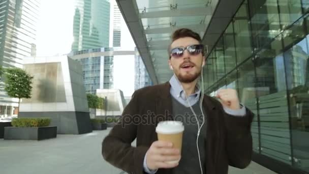 Beau homme écoutant de la musique dans les écouteurs smartphone et buvant du café sur papier, marchant et dansant — Video