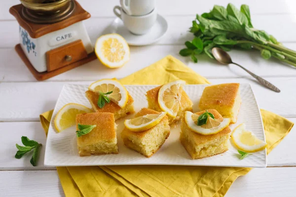 Square slices of lemon pie with slices of fresh lemon and mint leaves. A bunch of fresh mint in the corner on a wooden white background, a yellow cloth, half cut lemon, a manual coffee grinder, cups.