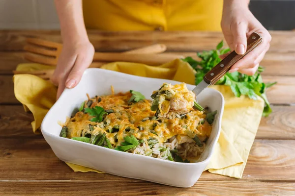 Baked chicken thigh fillet with rice, with cheese crust and soy sauce. Long-grain rice and green bean. White baking dish casserole on a wooden table, yellow fabric napkin, parsley leaves. Hold a dish