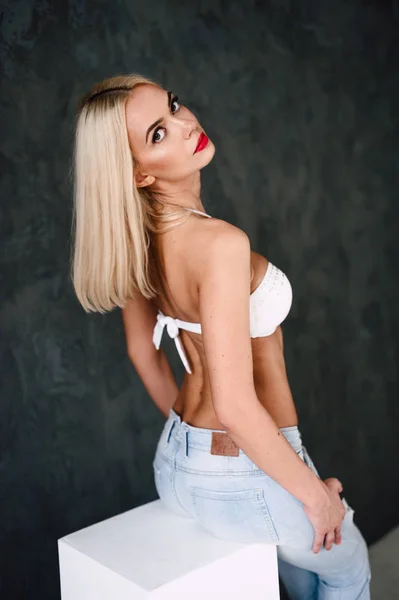 Гламурная молодая сексуальная блондинка в белом лифчике и джинсах с красными губами на темном фоне — стоковое фото