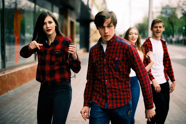 Four happy trendy teenage friends walking and dancing in the city, listening to the music with headphones, talking each other and smiling. Lifestyle, friendship and urban life concepts. — Stock Photo, Image