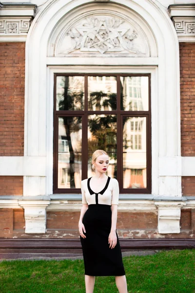 Mutlu moda sarışın Avrupa zarif kadın kırmızı dudakları ve beyaz ayakta yeşil çimenlerin üzerinde bina eski kırmızı tuğla, Cilt. Seksi kız açık havada. Resmi siyah beyaz elbiseli güzel kadın.